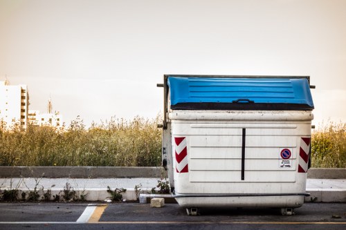 A large outdoor commercial rubbish container with a white body and a blue lid, positioned on a paved surface near a curb. The container is made of metal or durable plastic, with visible vertical ridges on its sides and red and white reflective chevron markings on the rear. It is set against a background of overgrown grass and shrubbery, with a partly cloudy sky above. The container appears to be used for waste collection or disposal, consistent with rubbish removal services in Hitchin or nearby areas. The scene suggests an urban or suburban environment, possibly on a parking lot or service area linked to property clearance and waste management activities. House Clearance Hitchin's services may include the removal and disposal of such waste containers as part of their house and home clearance offerings. The photograph captures the container in a neutral, functional setting, emphasizing its role in waste collection infrastructure.