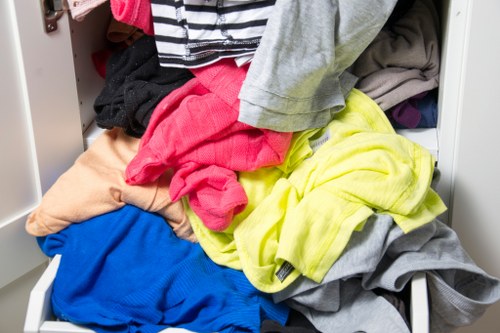 A close-up view of an overflowing white storage drawer filled with a variety of crumpled and unfolded clothing items, including brightly coloured garments such as a pink shirt, yellow hoodie, blue fabric, grey and beige pieces, and a black clothing item, with some textiles spilling over the edges. The surrounding environment suggests an indoor setting, potentially part of a house clearance process in Hitchin, where household textiles are being prepared for rubbish removal or disposal services provided by House Clearance Hitchin. The image emphasizes the disorderly state of laundry or textile waste awaiting collection, fitting within the context of waste management and house clearance activities in the local area.