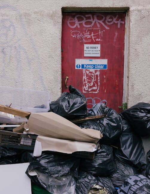 A residential alleyway scene featuring a red metal door with graffiti tags at the top, positioned within a beige, rough-textured wall. The door displays multiple signs, including a white sign with bla