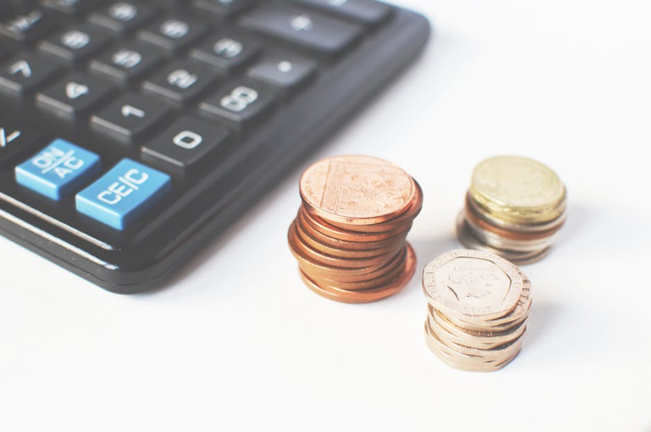 A close-up view of a black calculator with light blue function keys positioned on the left side of the image, with several neatly stacked piles of coins in front of it on a white surface. The coins in